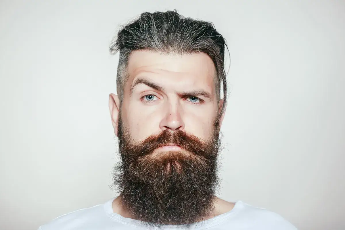 Hombre con barba larga y tupida, cabello rapado a los lados y peinado hacia atr&aacute;s. Sus ojos azules miran fijamente.