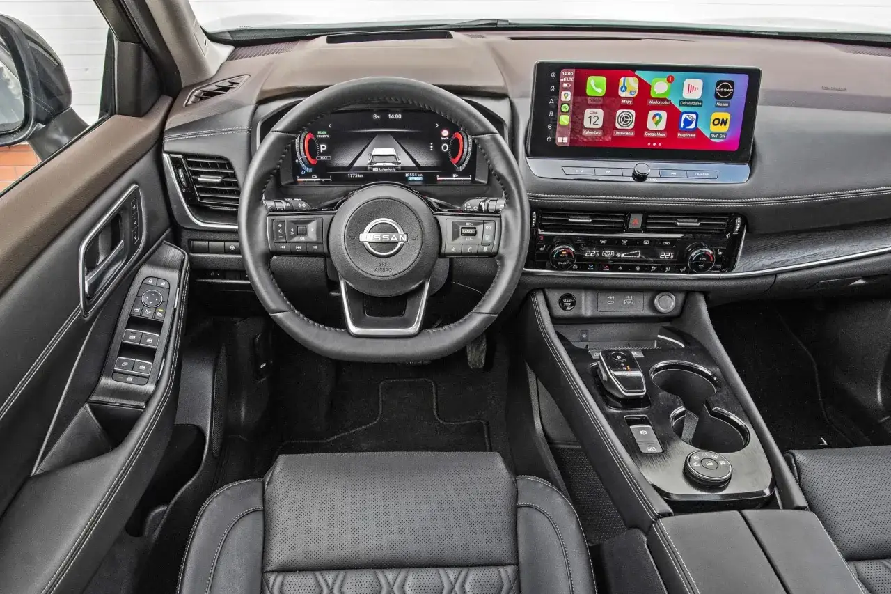 Wnętrze nowoczesnego Nissana X-Trail z cyfrowymi zegarami, dużym ekranem multimedialnym i skórzaną tapicerką.