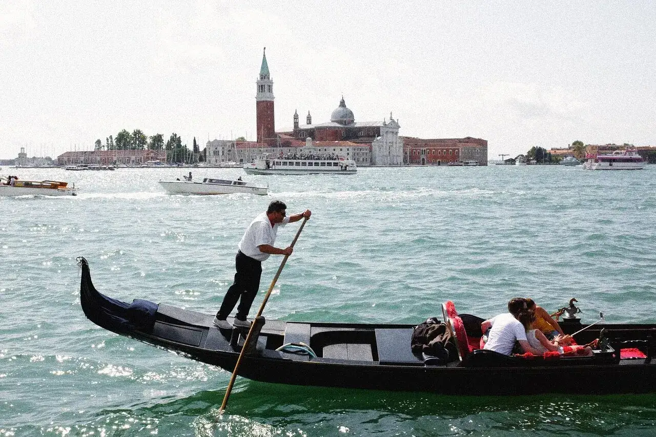 En gondolj&auml;r styr sin gondol med passagerare genom Venedigs kanaler, med kyrkor och palats i bakgrunden. En perfekt resa med t&aring;g till Venedig.