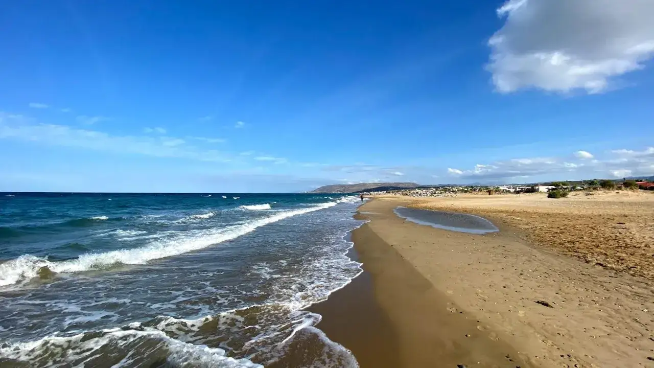 Spokojne fale obmywają piaszczysty brzeg Kavros beach. W oddali widać zabudowania i wzgórza.