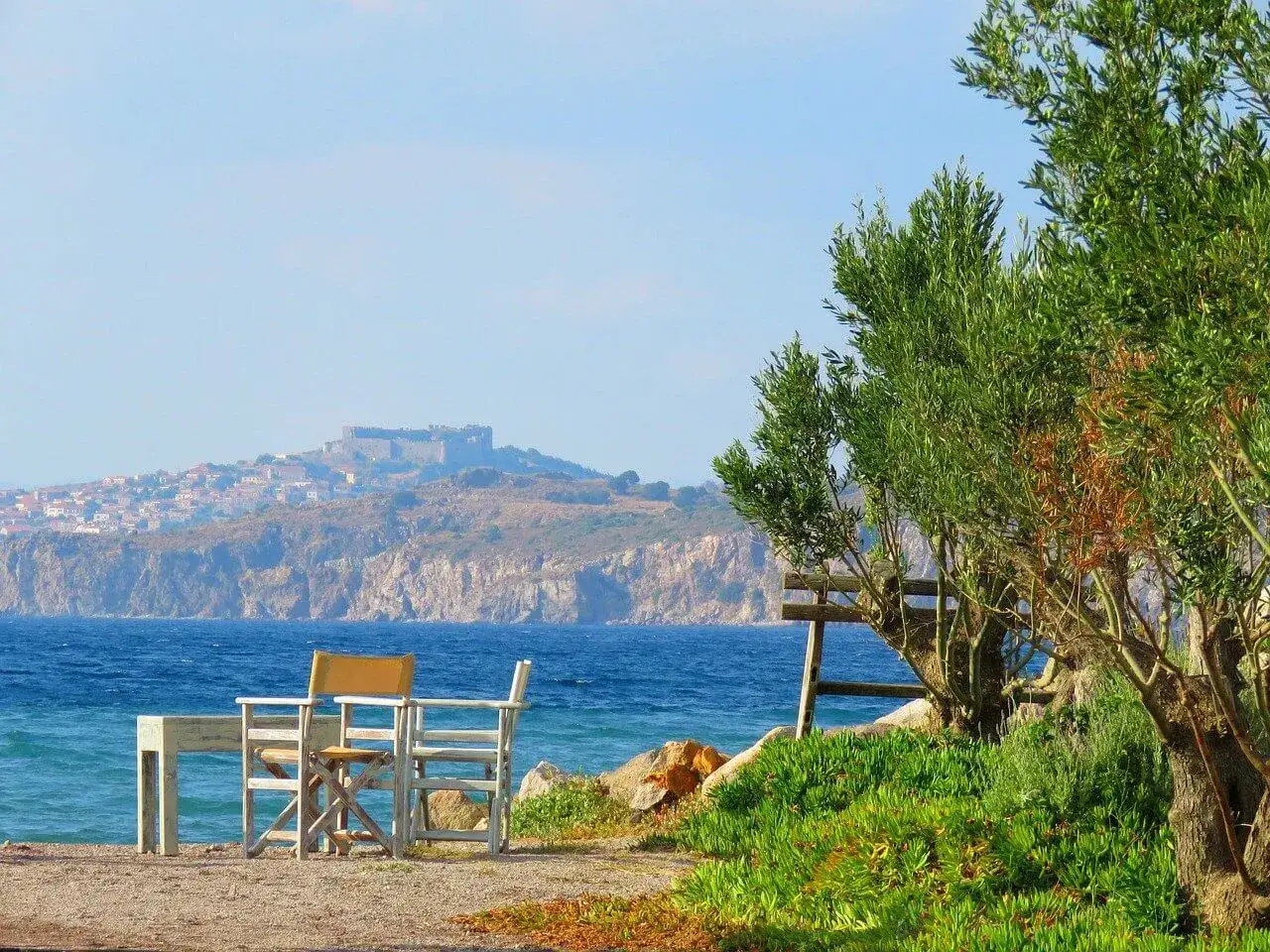 Piękna pogoda na Lesbos. St&oacute;ł i krzesła czekają na plaży, w tle widać zamek i miasteczko.