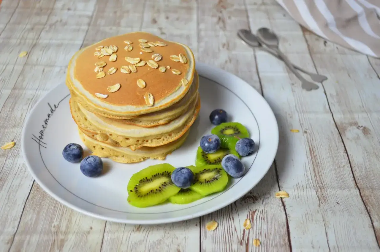 Pancake con albumi, guarniti con fiocchi d'avena, mirtilli e kiwi, su un piatto bianco con la scritta 