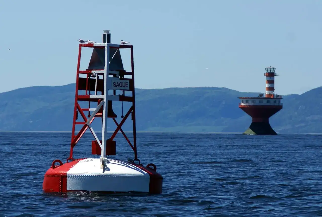 Boa rossa e bianca galleggia sull'acqua blu, con un faro a strisce in lontananza.
