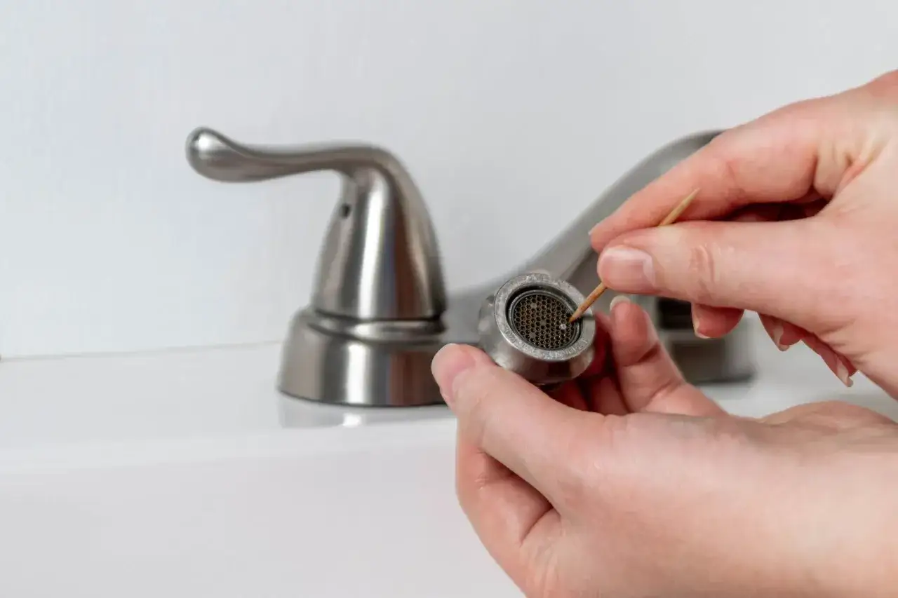 Hands carefully clean a faucet head with a toothpick, demonstrating how to clean faucet head to remove mineral deposits.