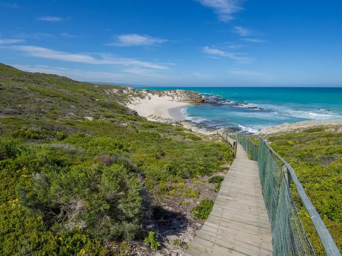 Holzsteg führt zum Strand im De Hoop Nature Reserve. Türkises Meer, grüne Hügel und blauer Himmel.