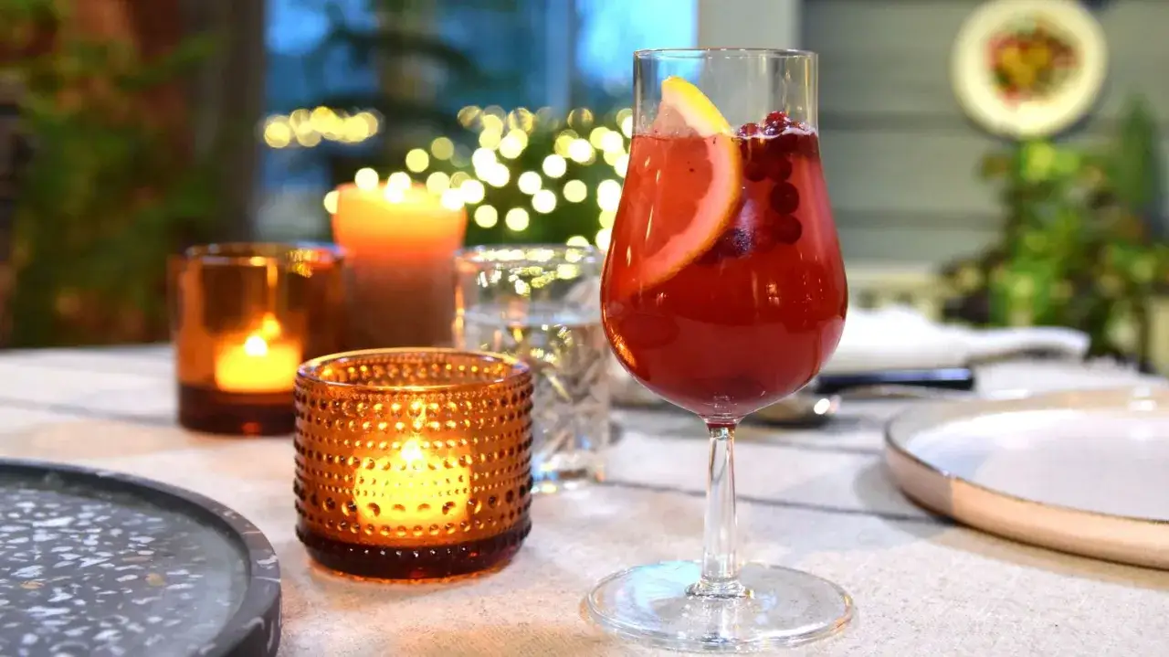 En glas med r&ouml;d dryck, garnerad med apelsinskiva och lingon. Ljus och juldekorationer i bakgrunden skapar en mysig st&auml;mning.