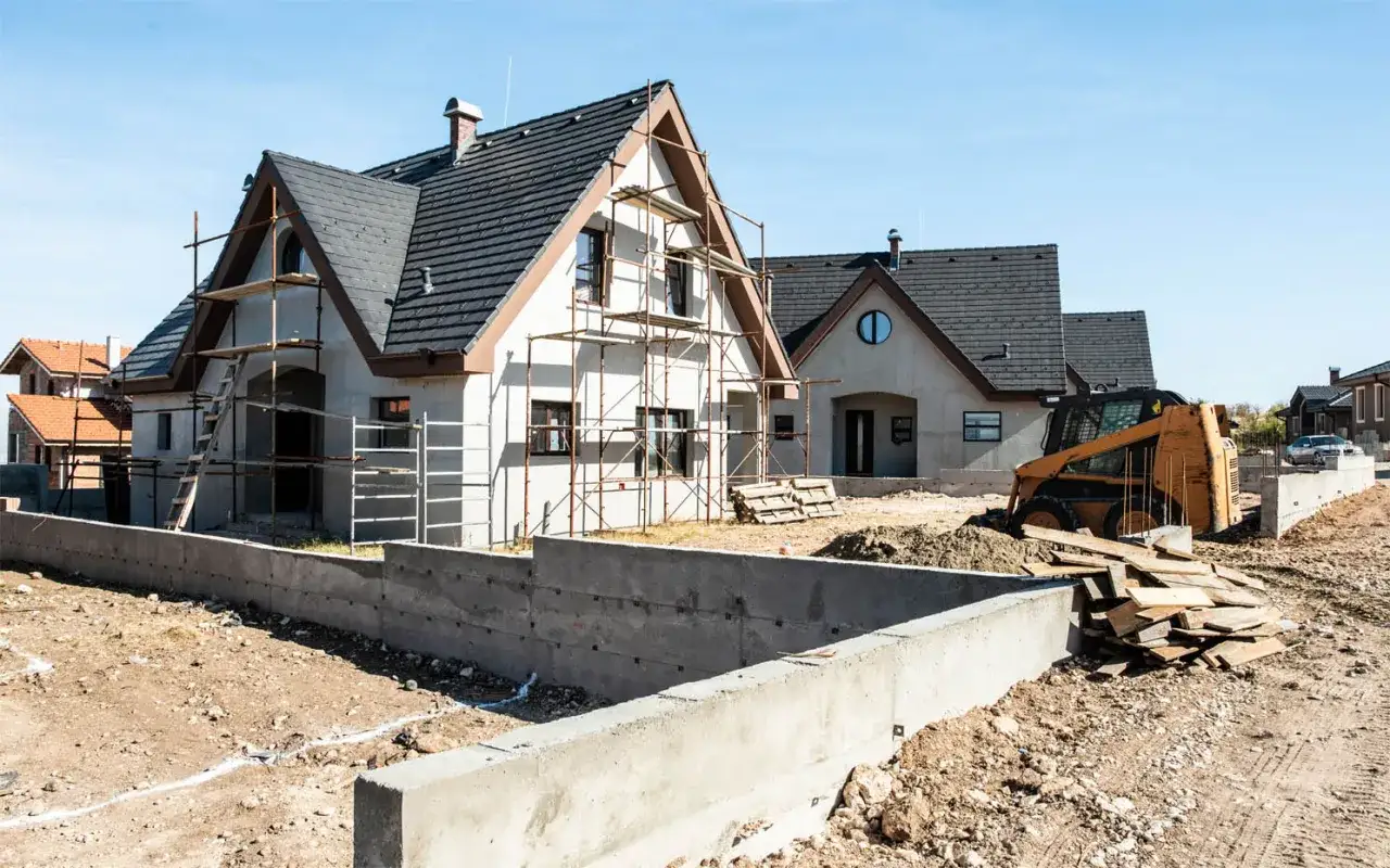 Budowa domów z bloczków betonowych. Na placu budowy widać domy w trakcie budowy, rusztowania, koparkę i materiały budowlane.