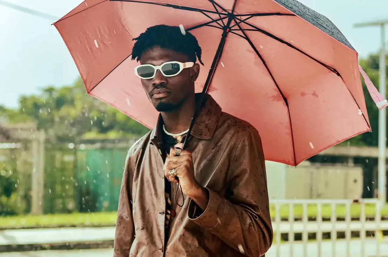 Un homme sous un parapluie rose, portant une veste en cuir marron, des lunettes blanches et un collier de perles. Il montre comment s'habiller quand il pleut et qu'il fait froid.