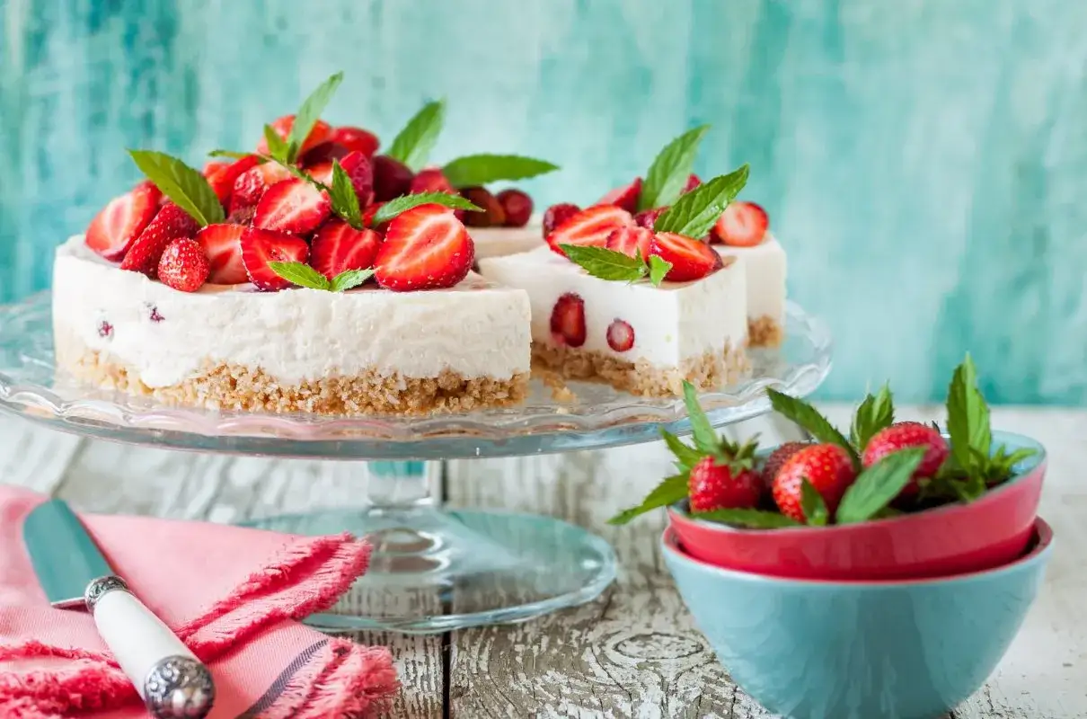 Szybkie ciasto z serkiem mascarpone, ozdobione świeżymi truskawkami i miętą, prezentuje się apetycznie na szklanej paterze.