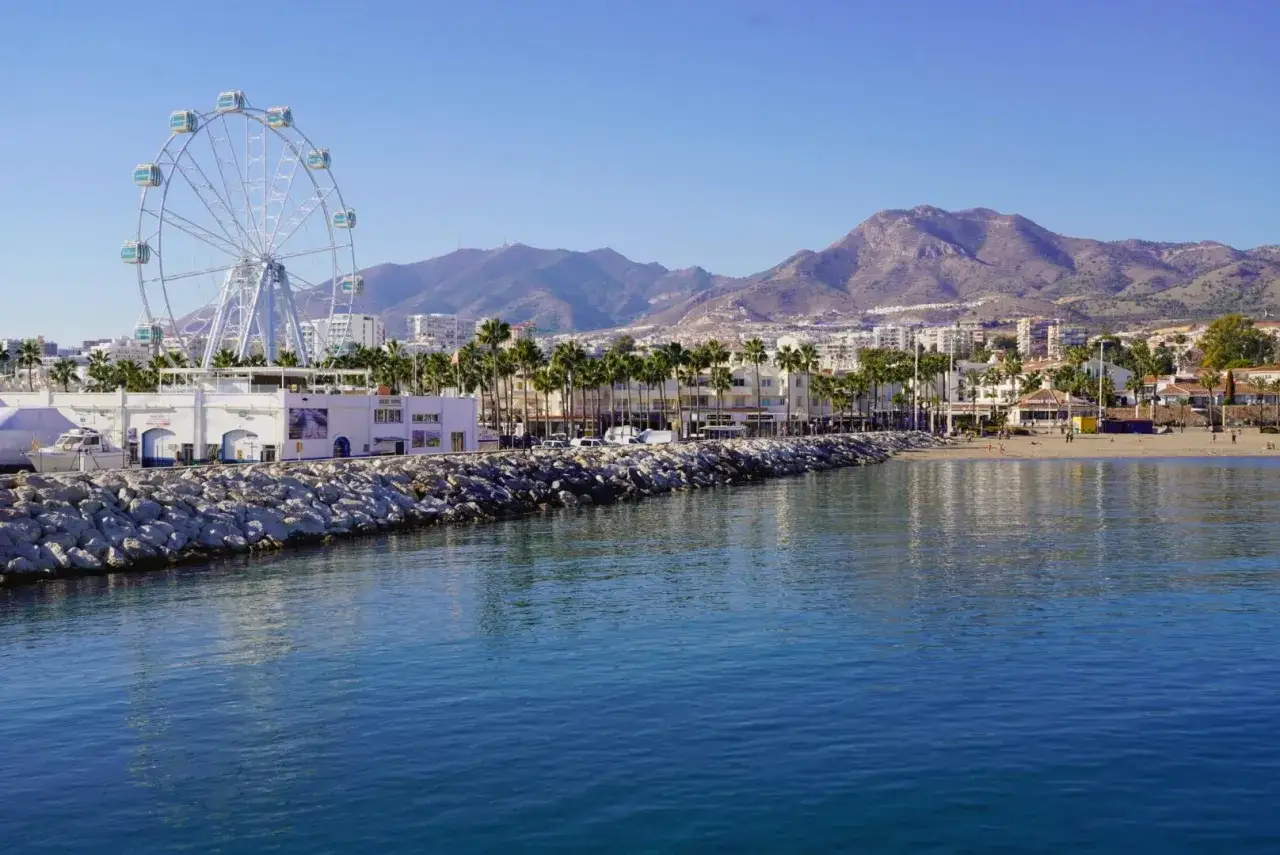 La playa fuente de la salud, con su noria y palmeras, se extiende bajo montañas.