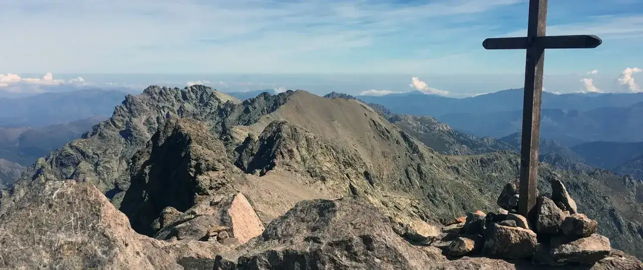 Croix en bois sur un sommet corse, dominant un paysage montagneux spectaculaire sous un ciel bleu et nuageux.