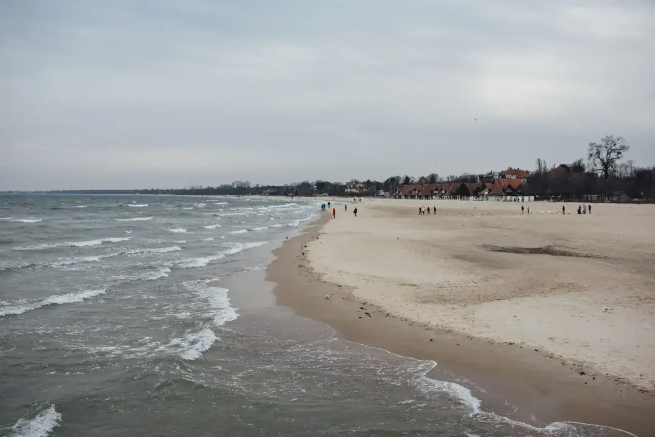 Plaża nad Bałtykiem, kilka os&oacute;b spaceruje, budynki w tle. Idealne miejsce na spokojne chwile, kiedy maj&oacute;wka.