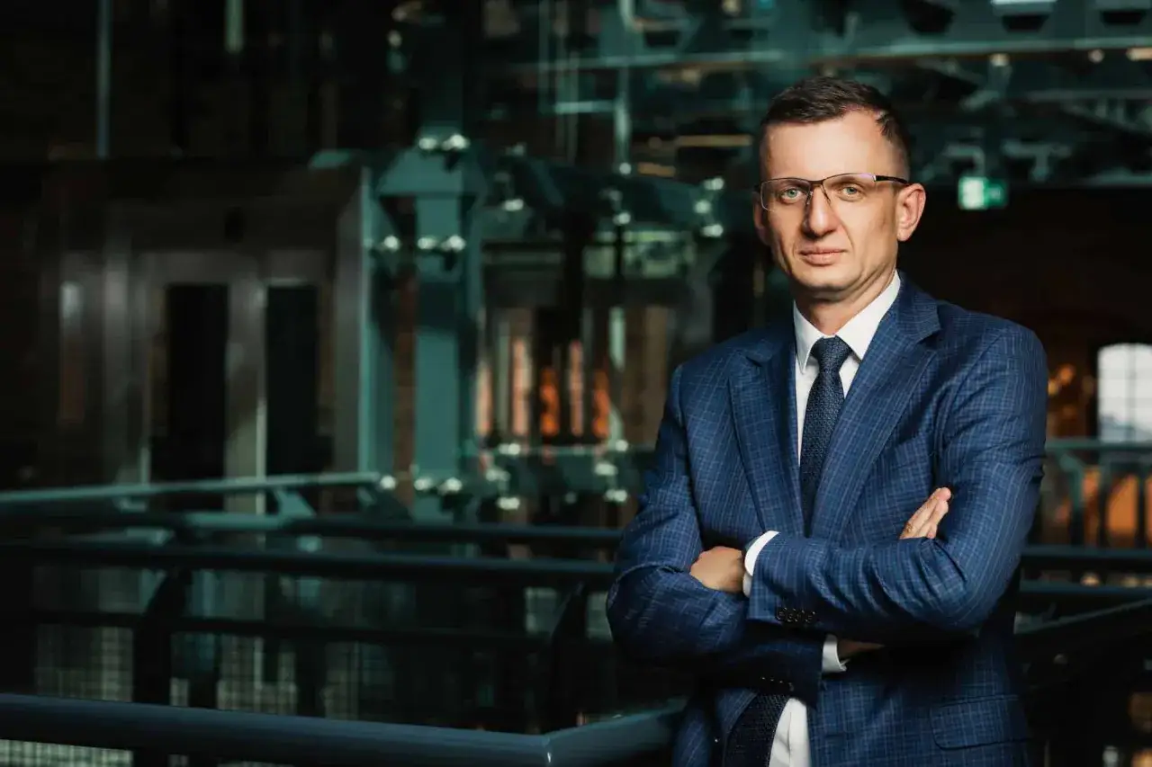 Mężczyzna w garniturze i okularach ze skrzyżowanymi rękami, na tle nowoczesnego wnętrza.