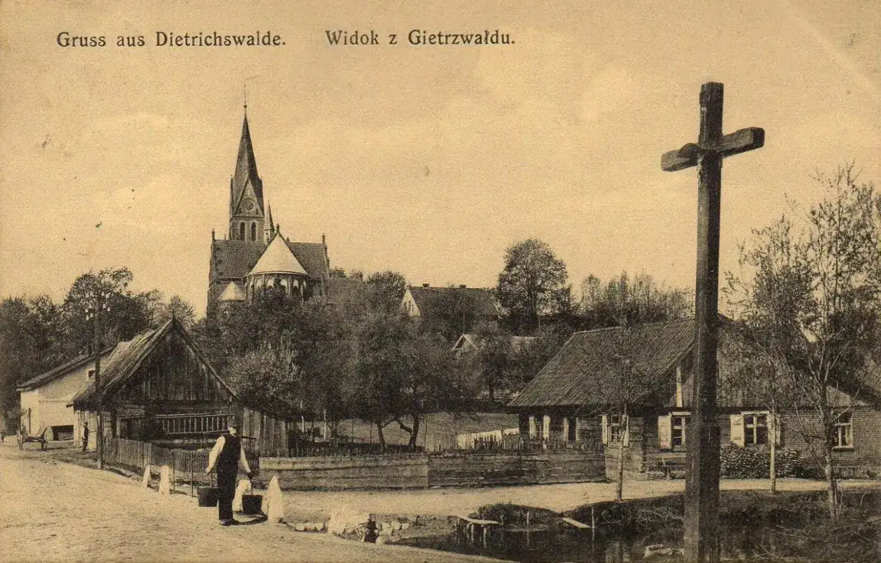 Widok z Gietrzwałdu: kościół, drewniane domy i krzyż. Mężczyzna niesie wiadra.