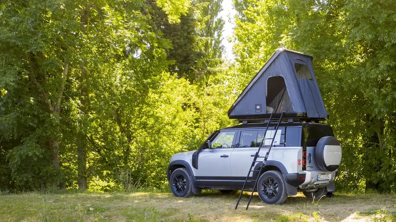 Srebrny Land Rover Defender z namiotem dachowym gotowy na spanie w samochodzie w Europie, zaparkowany w słonecznym lesie.
