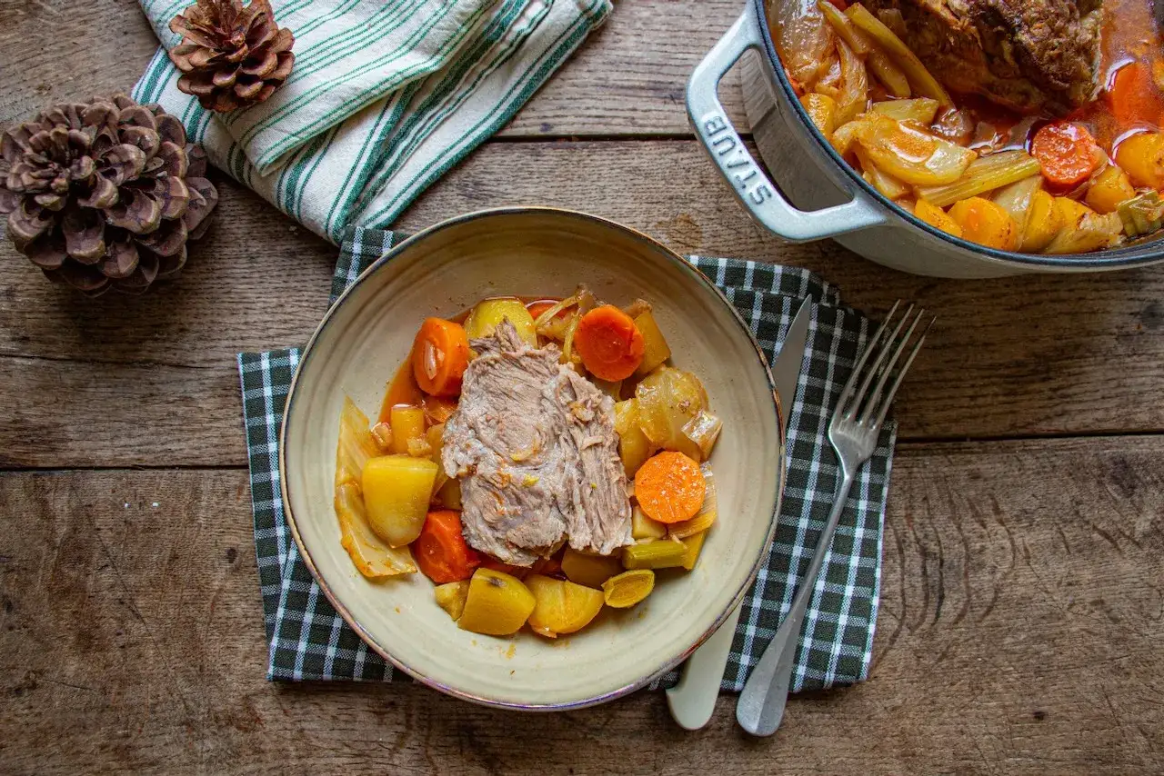 Un plat réconfortant de roti de porc cocotte, servi avec des carottes, pommes de terre et poireaux sur une table en bois rustique.