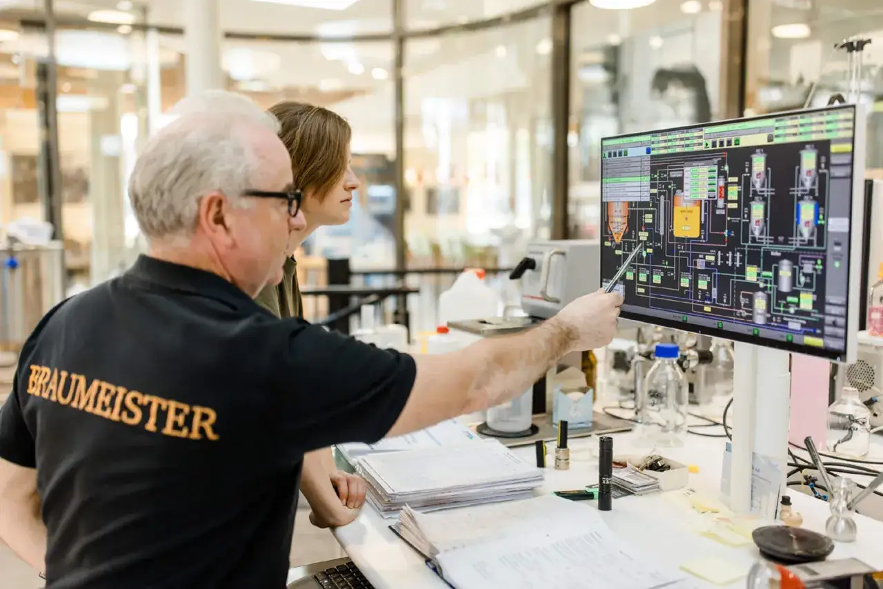 Erfahrener Braumeister erkl&auml;rt einem Kollegen die Prozesssteuerung auf einem Monitor. Ein Blick hinter die Kulissen von Braumeister Jobs.