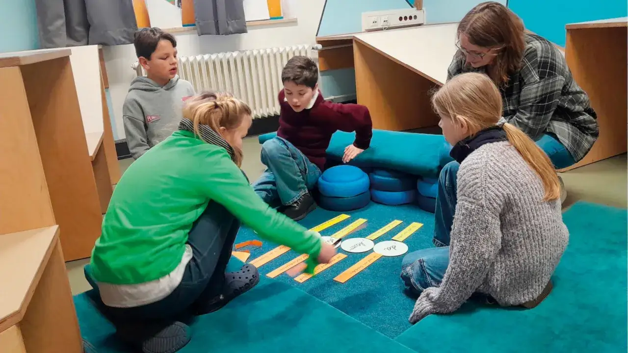 Kinder und eine Lehrerin bei einer Hospitation in der Schule, die auf dem Boden sitzen und mit Karten arbeiten.
