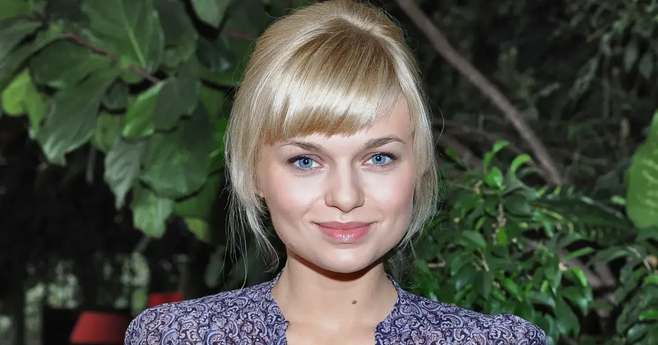 Uśmiechnięta aktorka na dobre i na złe z niebieskimi oczami i blond grzywką, na tle zielonych liści.