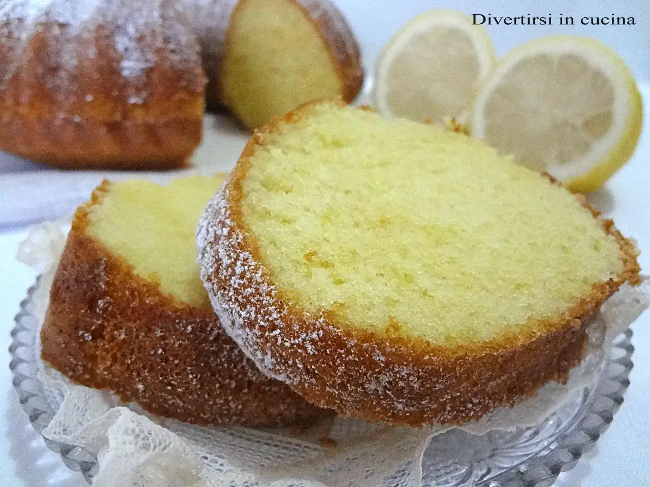 Fette di un soffice ciambellone al limone, spolverate di zucchero a velo, con spicchi di limone sullo sfondo.