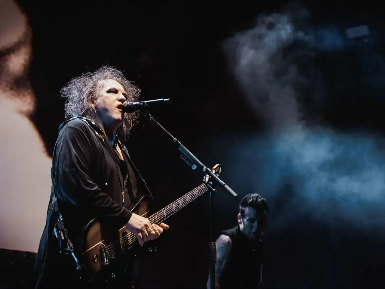 Robert Smith z The Cure Polska na scenie, w dymie i mroku, gra na gitarze.