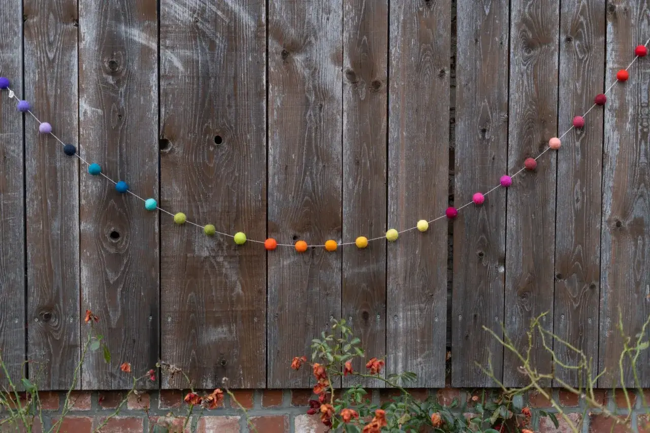Bunte Filzgirlande mit Kugeln in Regenbogenfarben, die sich &uuml;ber eine rustikale Holzlattenwand schl&auml;ngelt.