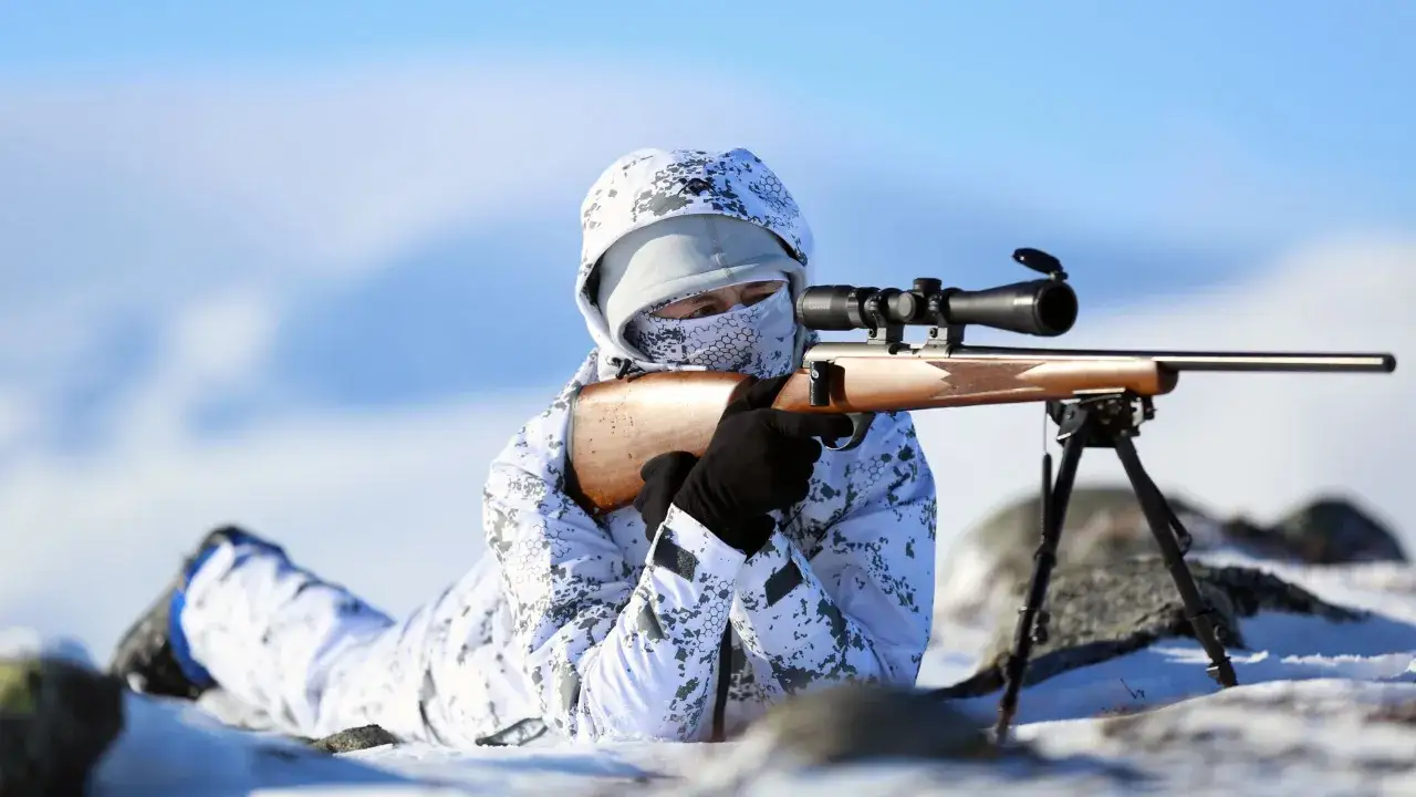 Person i kamouflerad vinterdräkt ligger i snön och siktar med ett gevär med kikarsikte. Kanske för att **snara ripa** i den karga svenska naturen.