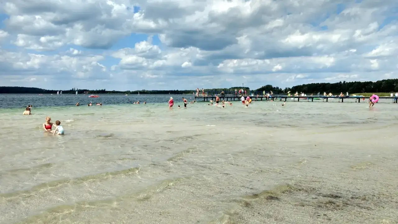 Ludzie kąpią się i relaksują nad Jeziorem Świętajno. W tle widać pomost i las.