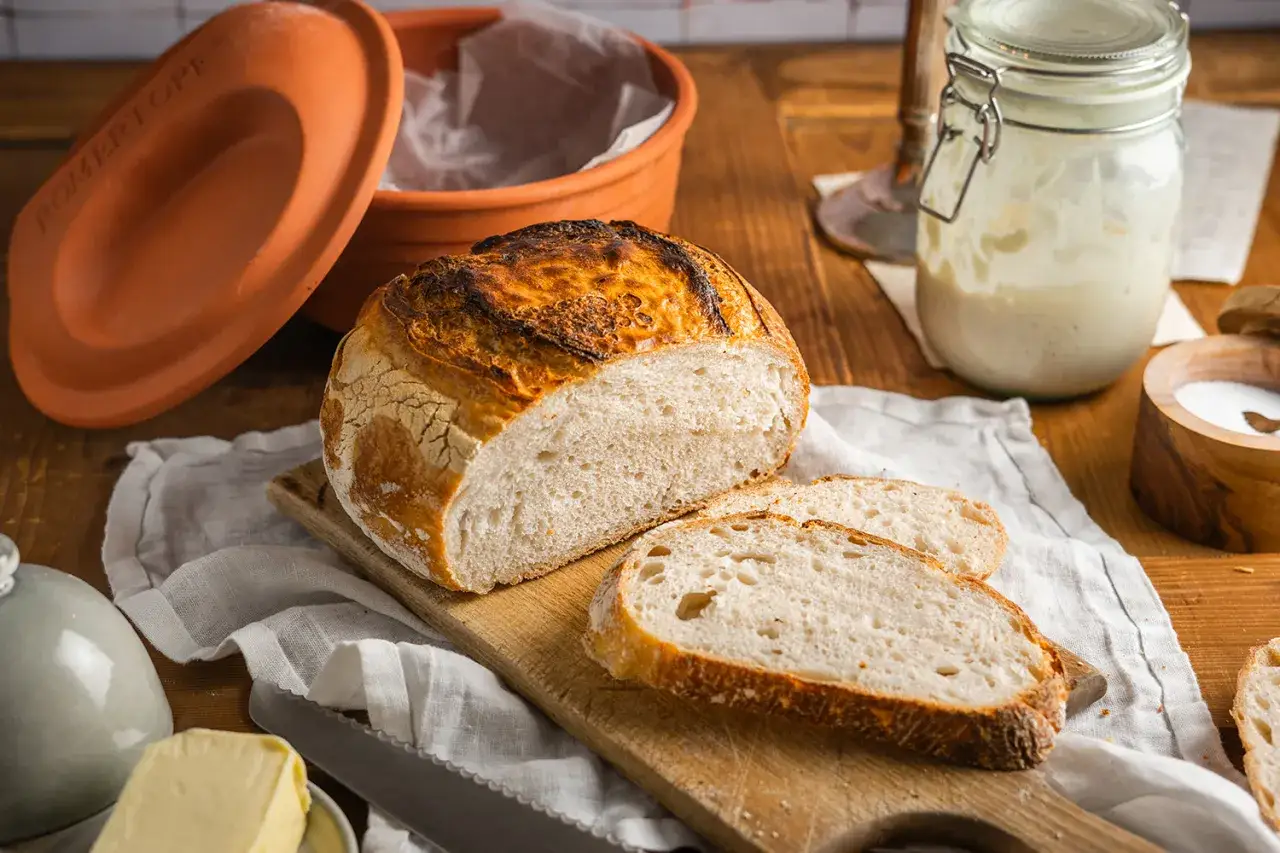 Frisch gebackenes **Sauerteig Weizen** Brot, aufgeschnitten auf einem Holzbrett, daneben ein Glas Sauerteig-Starter und ein Tontopf.