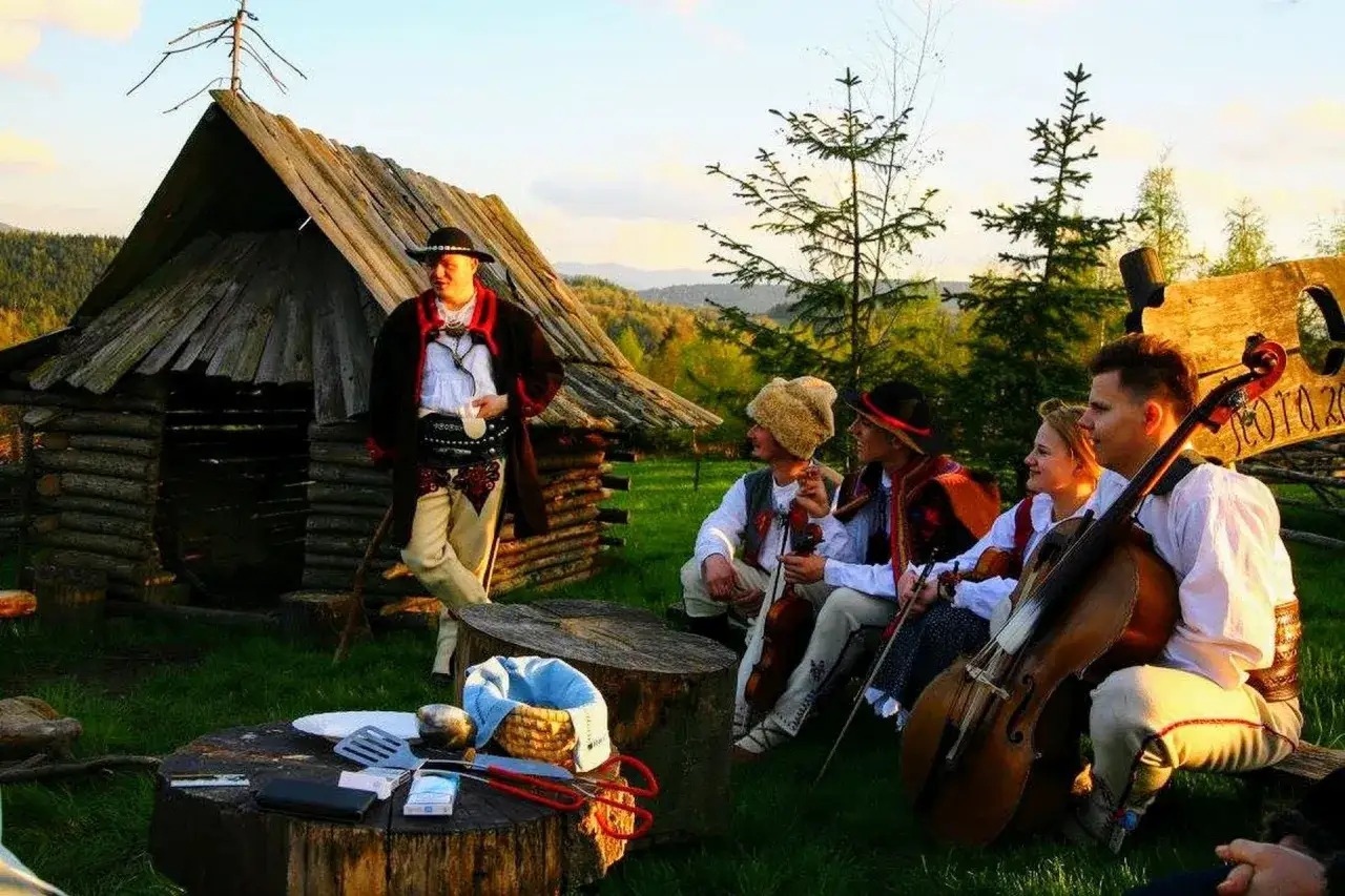 Muzycy w strojach góralskich grają przy bacówce. W tle drewniana chata i drzewa.