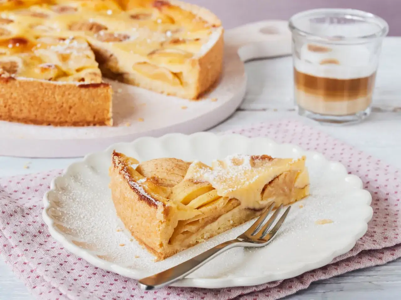 Ein Stück saftiger Apfelkuchen mit Guss, bestäubt mit Puderzucker, wartet auf dem Teller. Daneben ein Glas Latte Macchiato.