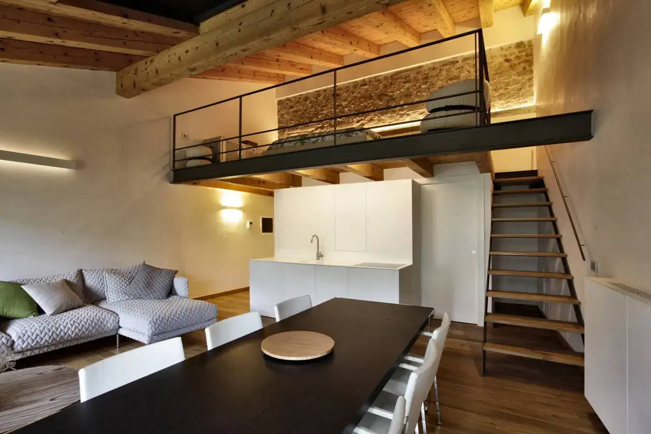 Modern living space with a loft bedroom accessible via stairs. The **soppalco soffitto basso** features a metal railing and a stone wall.