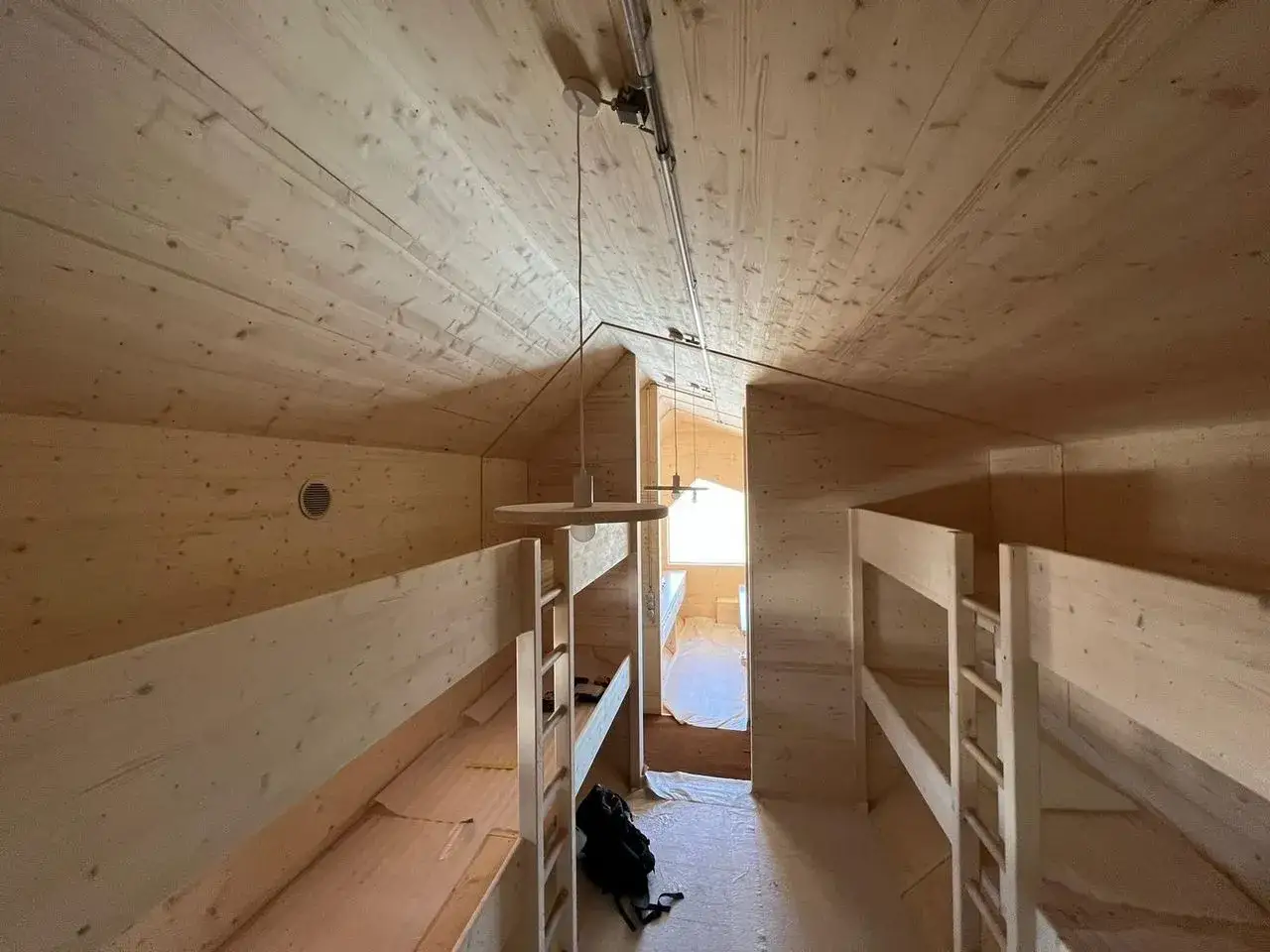 Interno di un bivacco in legno con letti a castello. Un sacco nero &egrave; appoggiato a terra.