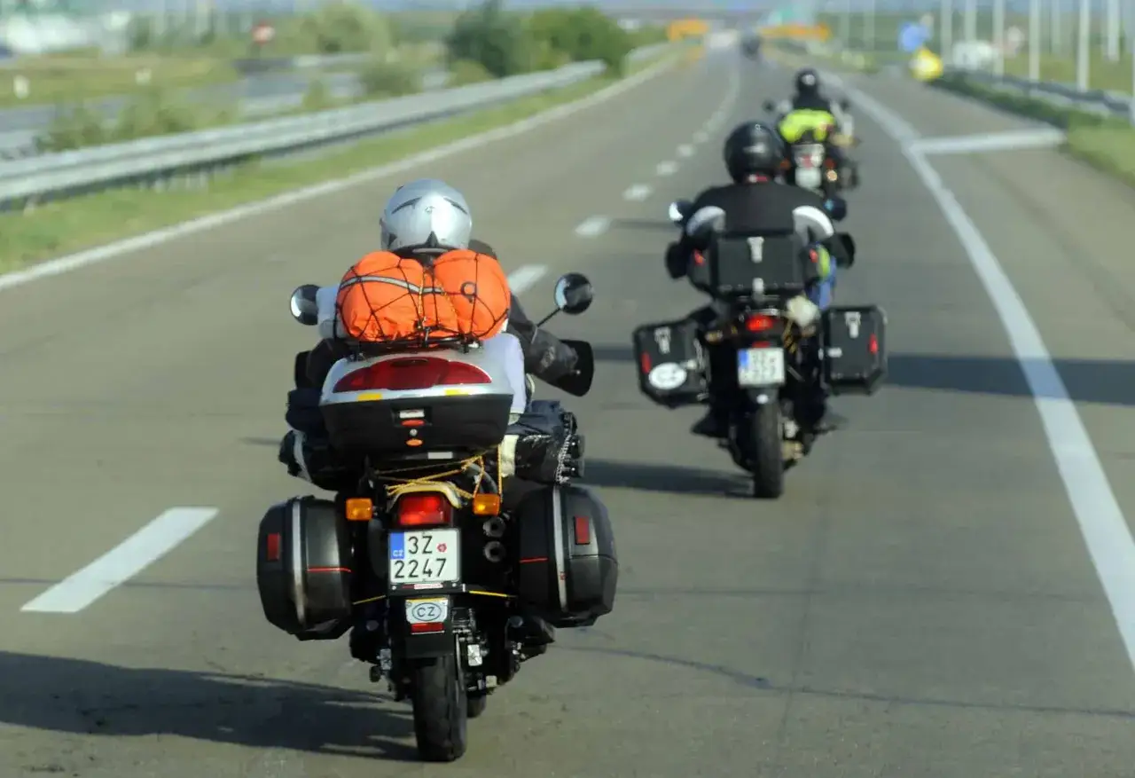 Motocykliści w kaskach i z bagażami jadą autostradą. Jeden z nich ma pomarańczowy bagaż na kufrze. Kategoria A2.