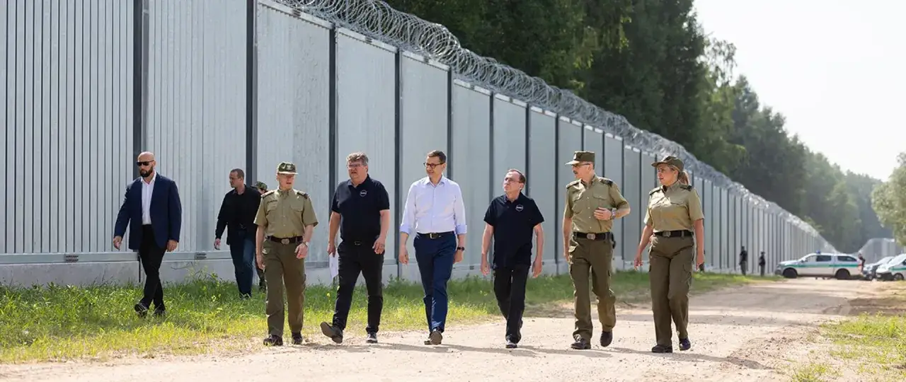 Grupa osób, w tym funkcjonariusze Straży Granicznej, idzie wzdłuż ogrodzenia na granicy polsko-białoruskiej.