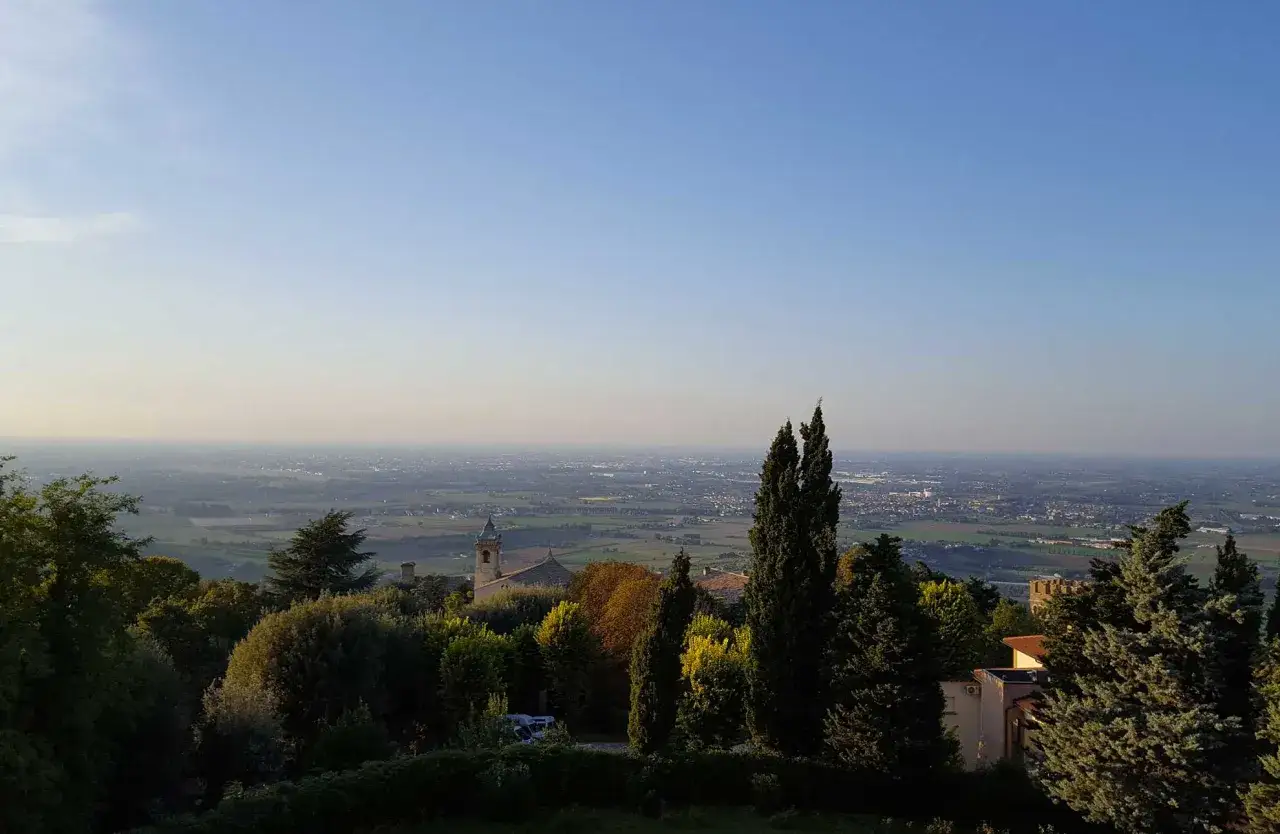 Vista panoramica dalla collina verso la pianura, con un campanile e alberi in primo piano. Si intravede la costa della riviera romagnola in lontananza.