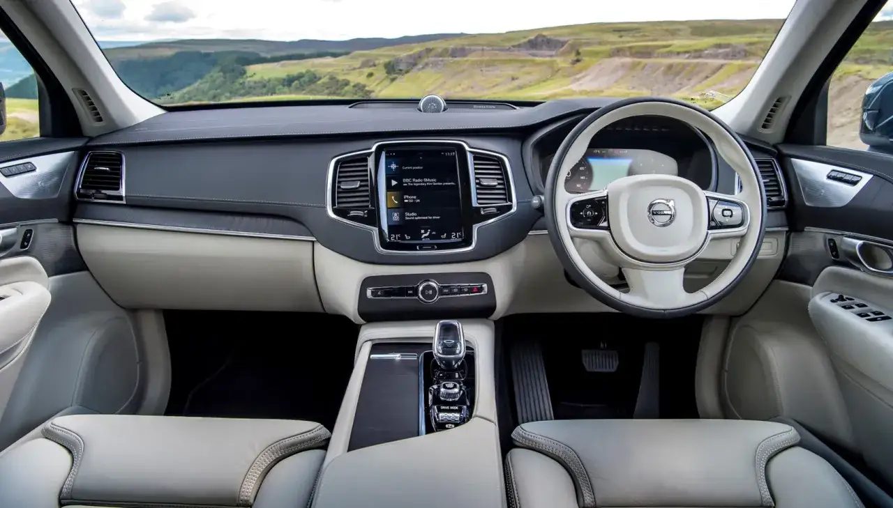 Wnętrze Volvo XC90 2020 z jasną tapicerką, dużym ekranem dotykowym i kierownicą. Widok na krajobraz za oknem.