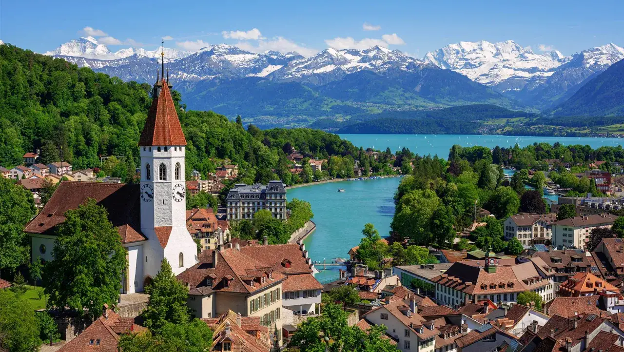 A white church with a red roof stands in a Swiss town, overlooking a turquoise lake and snow-capped mountains. This is one of nature Switzerland beautiful places.