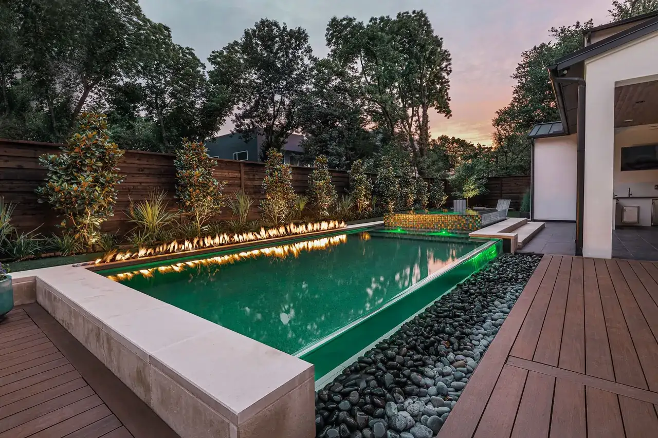 Moderne Poolumrandungen Ideen: Ein Pool mit grüner Beleuchtung, umgeben von Feuerstellen, Kies und Holzdeck.