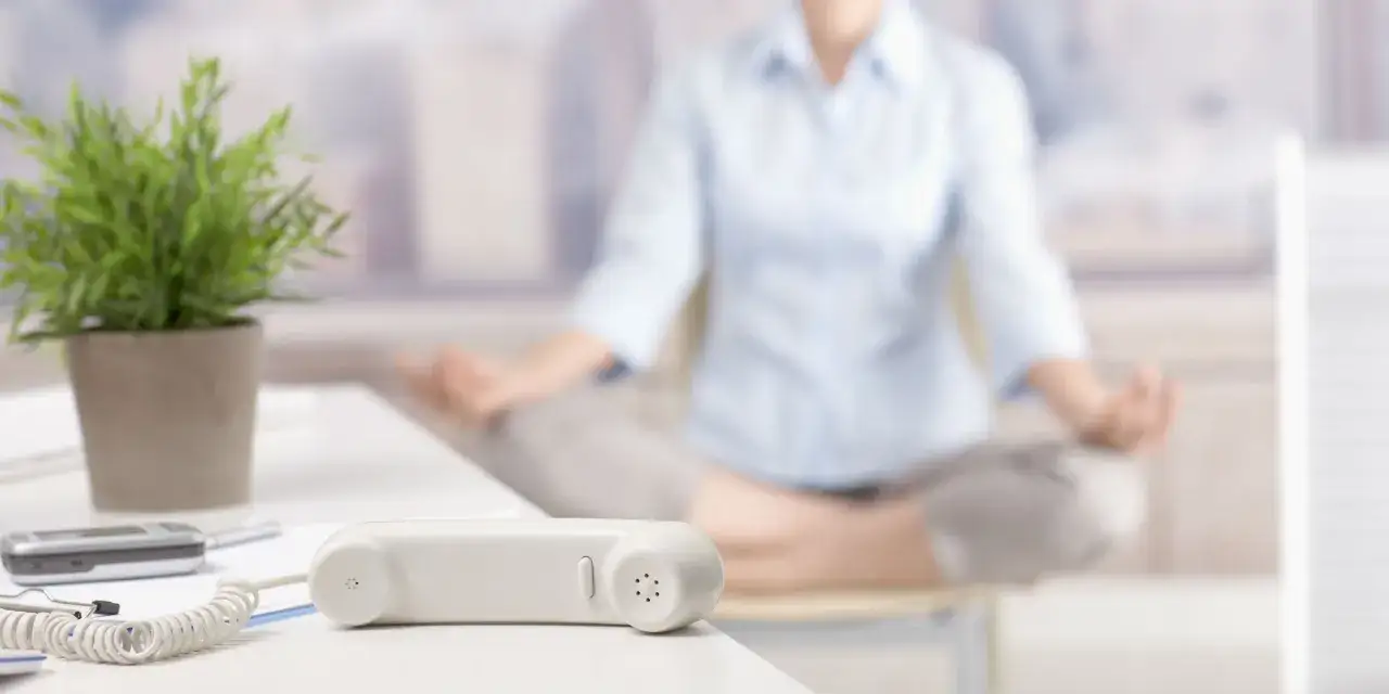 En person mediterar vid sitt skrivbord, en pausgympa p&aring; jobbet. En telefonlur och en v&auml;xt syns i f&ouml;rgrunden.