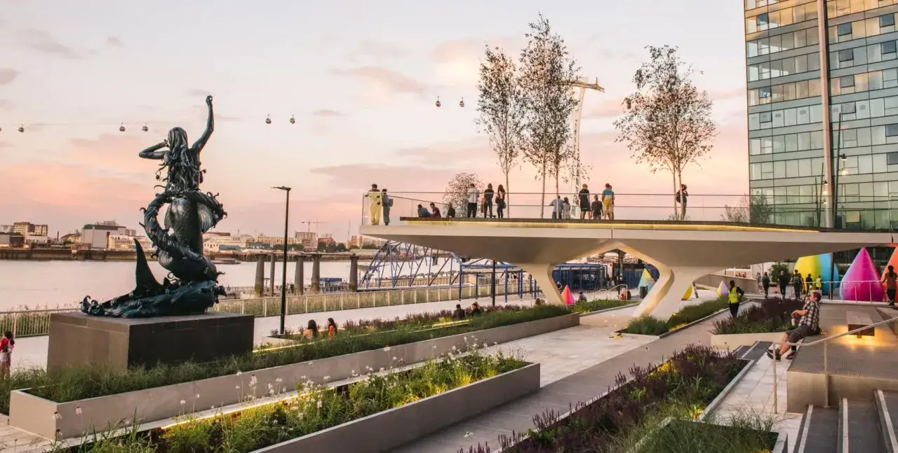 A striking mermaid sculpture, one of the area's famous sculptures, stands by the Thames at sunset, with people enjoying a modern walkway and colorful art installations.