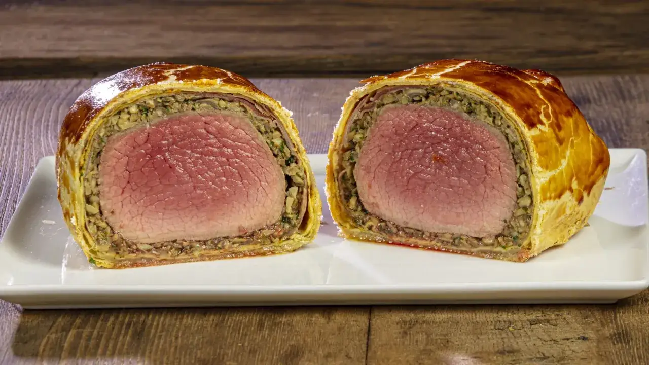 Un succulento filetto alla Wellington, tagliato a metà, rivela la carne rosata e il ricco ripieno di funghi, avvolti in una sfoglia dorata.