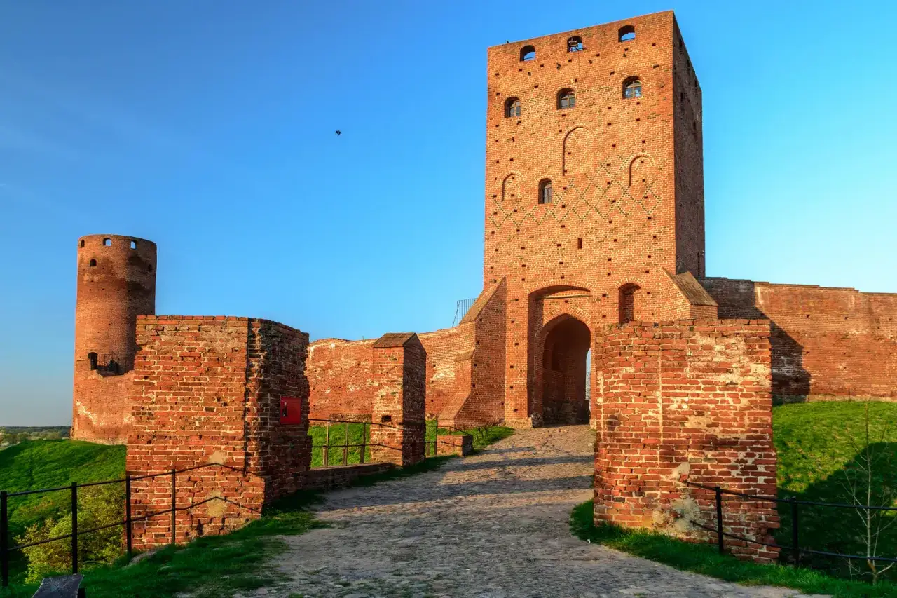 Ruiny zamku z cegły, z okrągłą wieżą i wysoką wieżą bramną. W oddali widać ptaka, jakby węgiełek lalka.