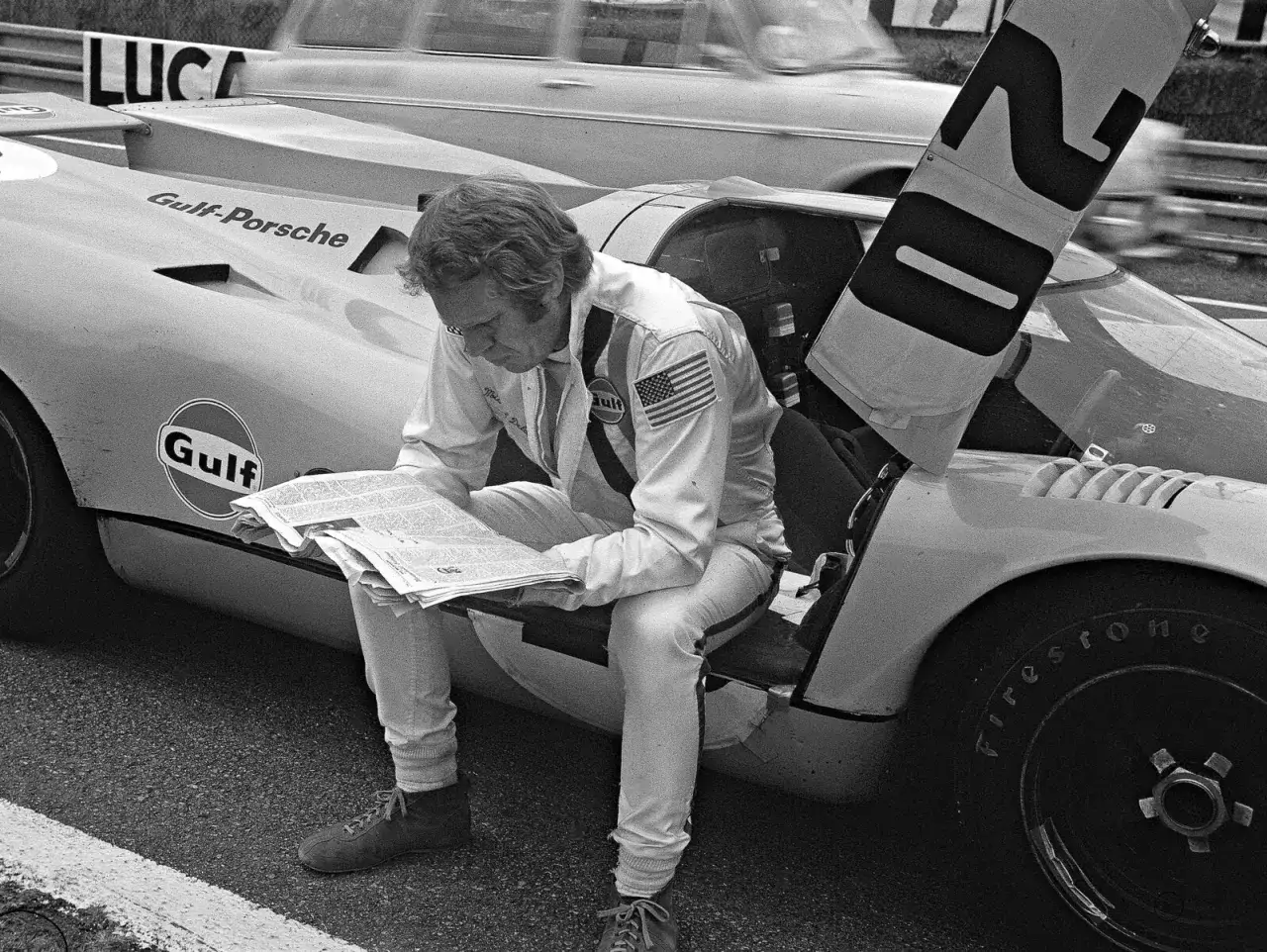 Steve McQueen w kombinezonie Gulf czyta gazetę, siedząc na swoim samochodzie wyścigowym Gulf-Porsche.