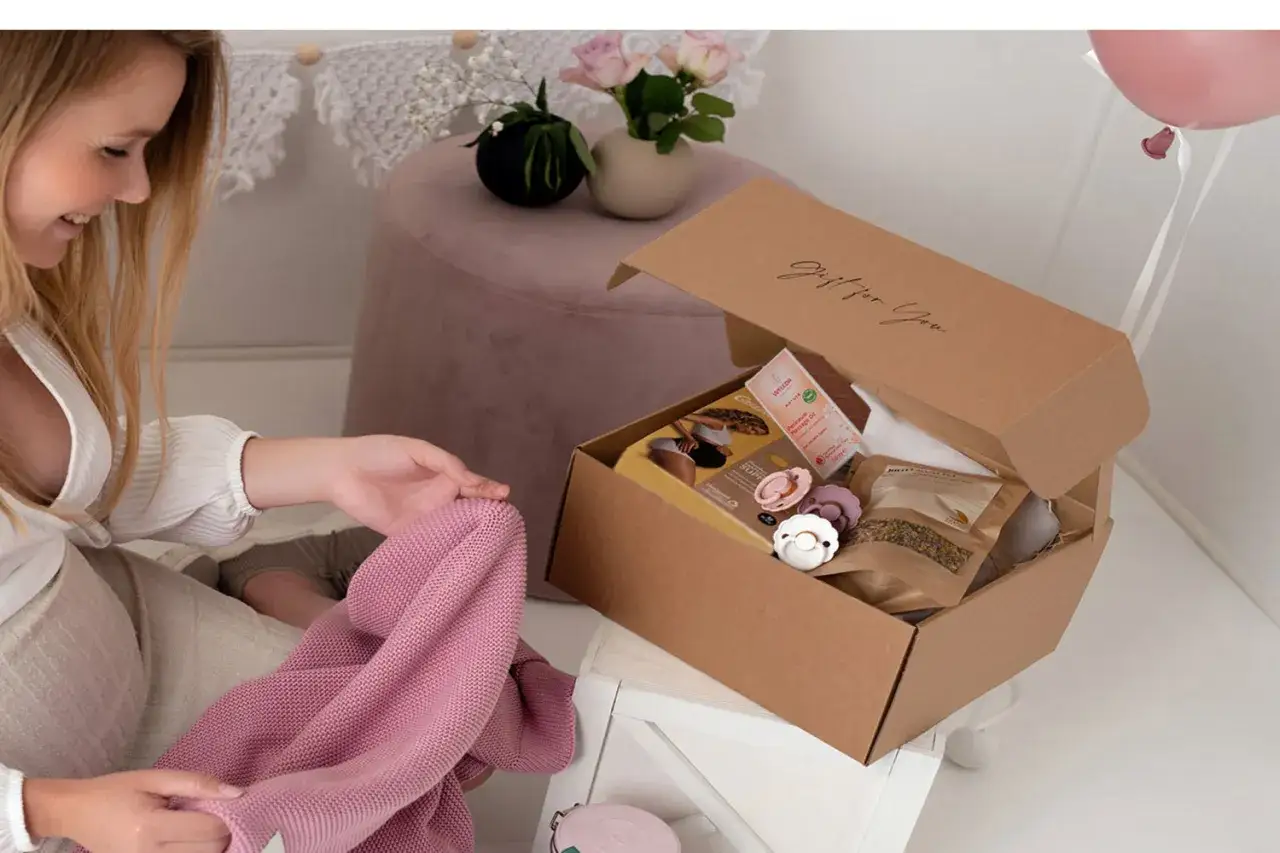 En kvinna &ouml;ppnar en nyf&ouml;dd present box med en mjuk rosa filt. L&aring;dan inneh&aring;ller nappar, olja och te.