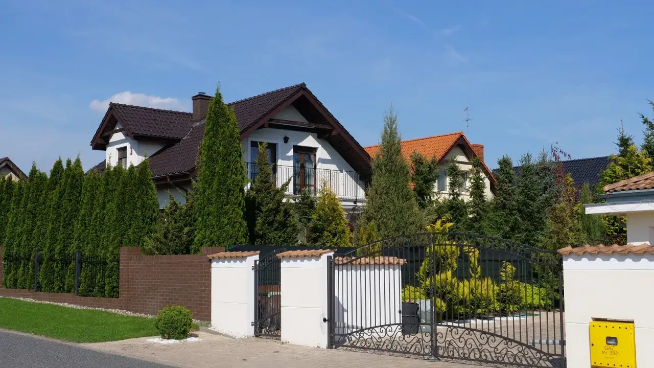 Żółta skrzynka gazowa w elewacji budynku, obok ozdobnej bramy i zielonych żywopłotów.