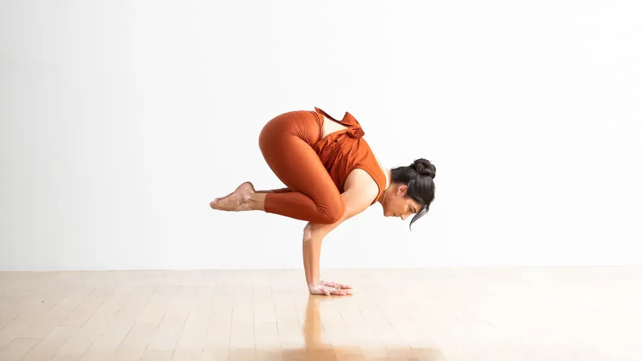 Mujer en la postura del cuervo yoga, con las rodillas dobladas sobre los brazos y el cuerpo en equilibrio.
