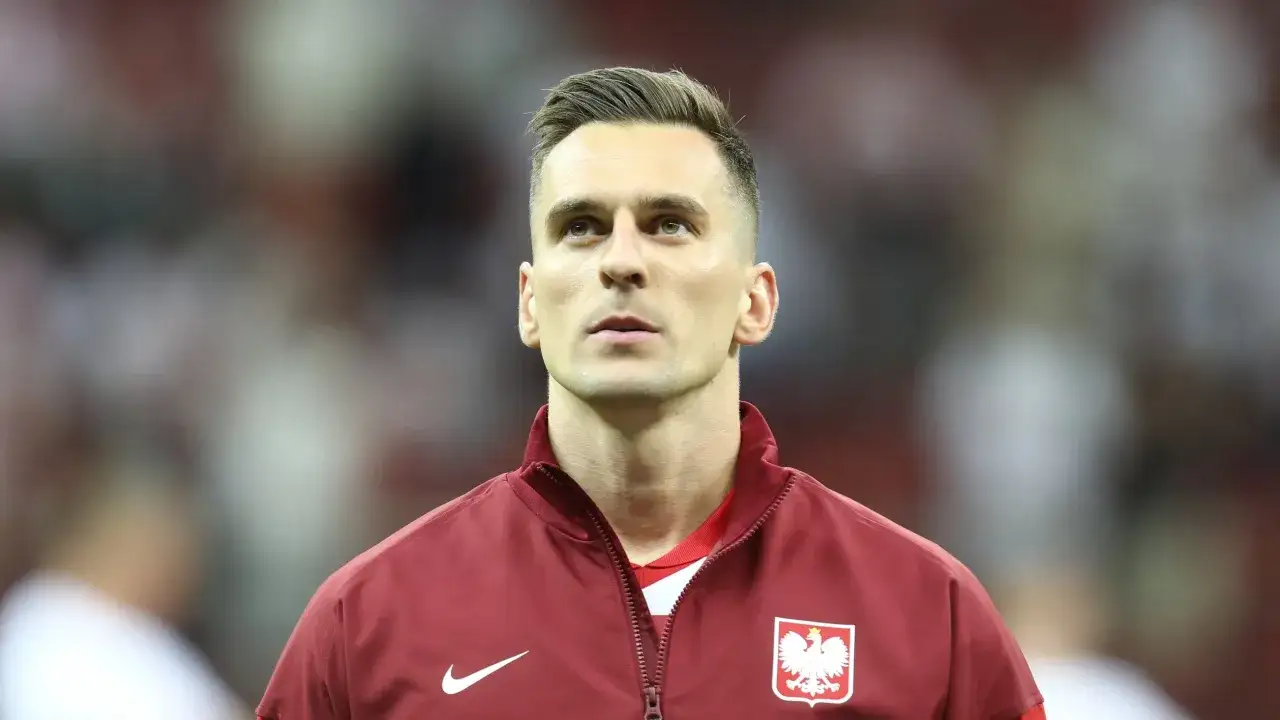 Piłkarz Milik w bordowej kurtce z orłem na piersi, gotowy do gry.