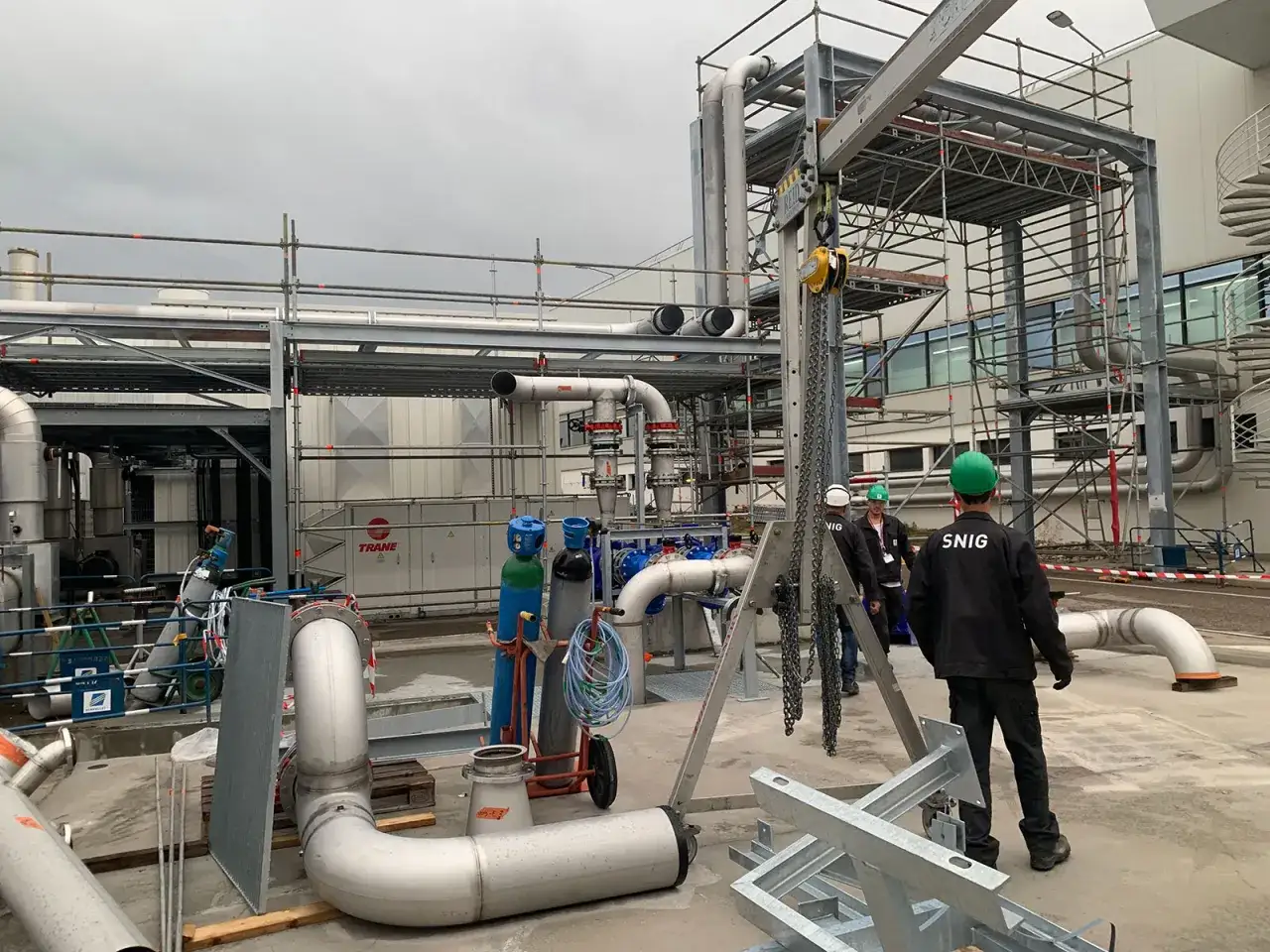Chantiers industriels avec tuyauterie, &eacute;chafaudages et personnel portant des casques. Un chariot &eacute;l&eacute;vateur est visible, ainsi que des bouteilles de gaz. Le logo SNIG est pr&eacute;sent sur un v&ecirc;tement.
