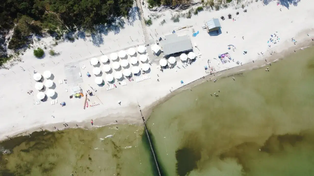 Plaża z białymi parasolami i leżakami, ludzie kąpią się w morzu. Idealne miejsce na wakacje last minute.